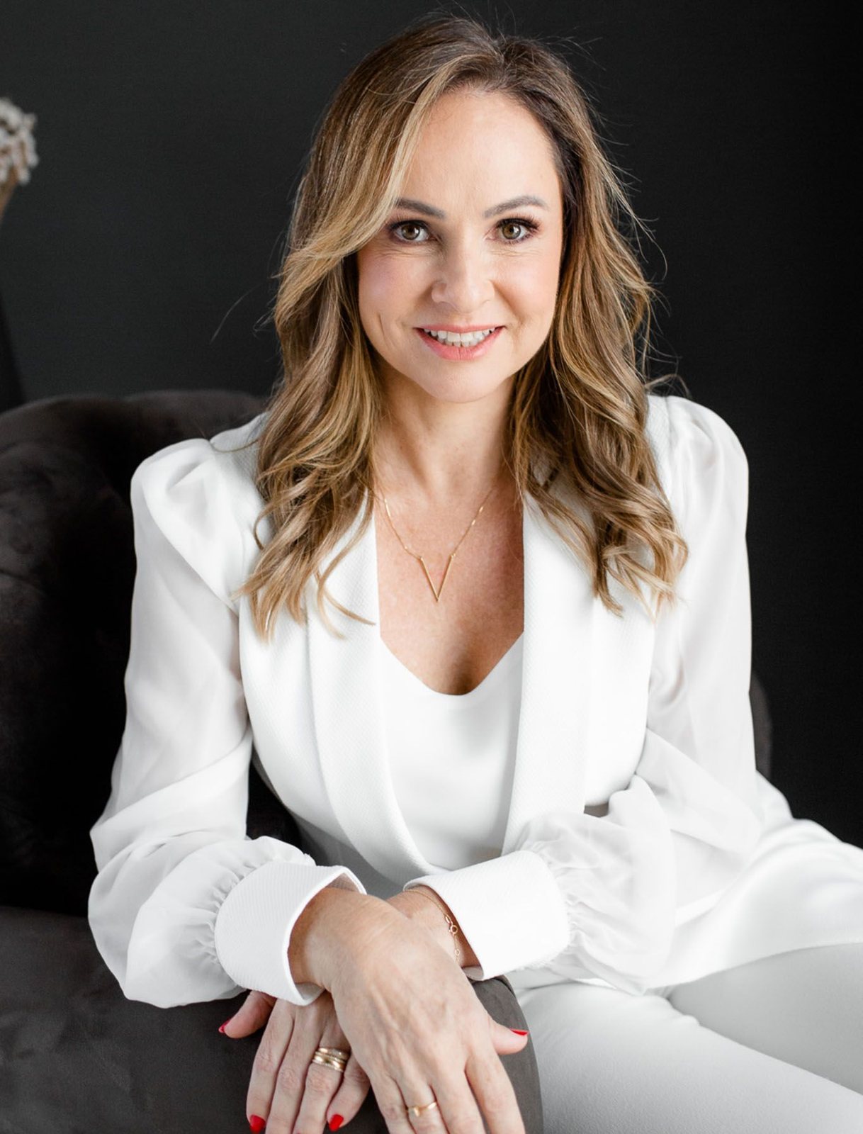 Dra. Alessandra Nalin sentada, de blazer branco, sorrindo, sobre fundo escuro